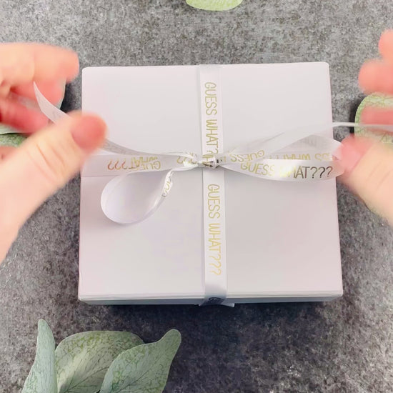 Unboxing a white box tied with white ribbon. In gold text printed on the ribbon it says "Guess What???". The box opens to reveal baby booties with a wooden tag attached, saying "You're going to be an Auntie", perfect for a pregnancy announcement